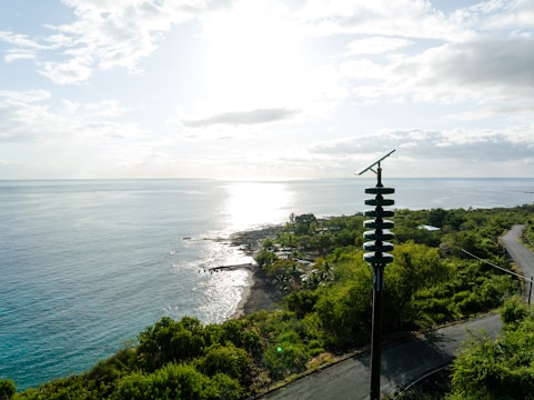 A scenic coastal view with the sun reflecting on the ocean, surrounded by lush green vegetation. A tall pole with solar panels is prominently visible in the foreground. The coastline features a rocky shore and calm waters.