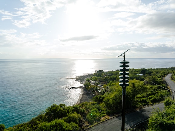 A scenic coastal view with the sun reflecting on the ocean, surrounded by lush green vegetation. A tall pole with solar panels is prominently visible in the foreground. The coastline features a rocky shore and calm waters.