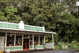 A quaint storefront with a green and white exterior situated in a lush, wooded area. The building features large windows displaying various items, suggesting it may be a shop. A red door is prominently centered, and a small white fence lines the side. The surrounding foliage is dense and vibrant, adding to the serene atmosphere.