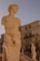 A close-up view of a large marble statue depicting a figure with a muscular build and curly hair. In the background, several other statues are visible along a staircase, and there is an old, weathered building with balconies and shuttered windows under a clear sky.