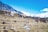 A mountainous landscape showcases traditional stone houses nestled on terraced slopes. The village is surrounded by snow-capped mountains under a bright blue sky. Colorful prayer flags add vibrancy to the rustic scenery, and there are people walking along a path among the terraces.