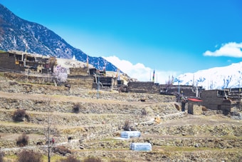 A mountainous landscape showcases traditional stone houses nestled on terraced slopes. The village is surrounded by snow-capped mountains under a bright blue sky. Colorful prayer flags add vibrancy to the rustic scenery, and there are people walking along a path among the terraces.