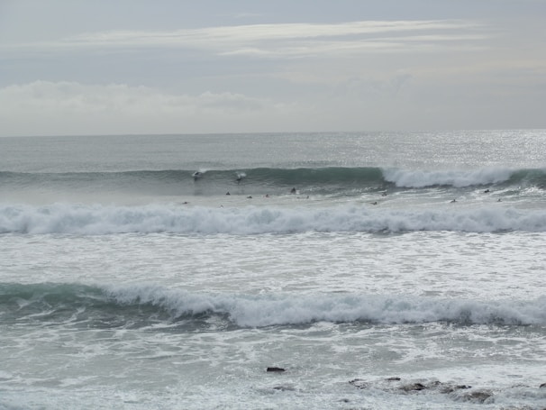 Large ocean waves roll towards the shore, with several surfers positioned to catch them. The sky is overcast, casting a gray tone over the scene, and the water appears turbulent with white foam.