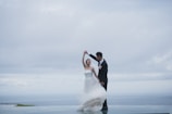 A couple is dancing gracefully on a rooftop or elevated platform with an expansive view of the ocean and a cloudy sky. The woman is wearing a flowing white wedding gown, and the man is dressed in a formal black suit with a boutonnière.