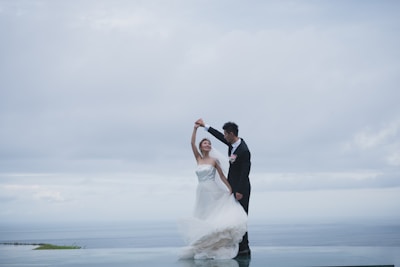 A couple is dancing gracefully on a rooftop or elevated platform with an expansive view of the ocean and a cloudy sky. The woman is wearing a flowing white wedding gown, and the man is dressed in a formal black suit with a boutonnière.