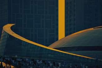 A modern architectural scene with a focus on a large, curved structure featuring glass surfaces. The building is contrasted by a bright yellow vertical element that stands out against the dark, textured background of other architectural elements.