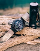 A rugged digital wristwatch with a fabric strap is resting on textured tree bark, accompanied by a black cylindrical flashlight and a multitool. The background is blurred, suggesting an outdoor setting.
