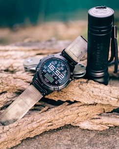 A rugged digital wristwatch with a fabric strap is resting on textured tree bark, accompanied by a black cylindrical flashlight and a multitool. The background is blurred, suggesting an outdoor setting.