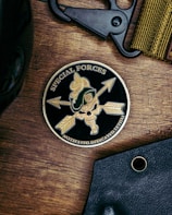 A round emblem featuring the words 'Special Forces' and 'Motivated. Dedicated. Lethal.' surrounds a design with arrows, a skull, and a green military helmet with a gold crest. The emblem is on a wooden surface, accompanied by a black tool and a fabric band.