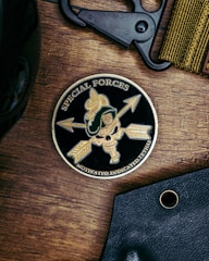 A round emblem featuring the words 'Special Forces' and 'Motivated. Dedicated. Lethal.' surrounds a design with arrows, a skull, and a green military helmet with a gold crest. The emblem is on a wooden surface, accompanied by a black tool and a fabric band.