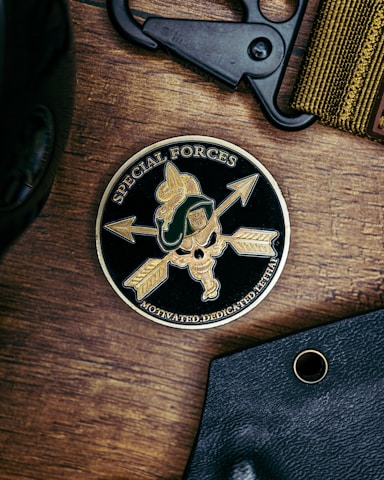 A round emblem featuring the words 'Special Forces' and 'Motivated. Dedicated. Lethal.' surrounds a design with arrows, a skull, and a green military helmet with a gold crest. The emblem is on a wooden surface, accompanied by a black tool and a fabric band.