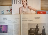 Several newspapers and magazines are arranged on a wooden surface. One magazine features a portrait of a woman with wet hair in a white top. Below are pages from a German newspaper with articles and images, including one of a man in a suit. The Economist magazine is partially visible on the right.