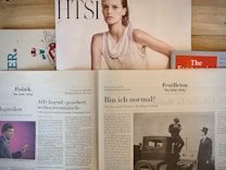 Several newspapers and magazines are arranged on a wooden surface. One magazine features a portrait of a woman with wet hair in a white top. Below are pages from a German newspaper with articles and images, including one of a man in a suit. The Economist magazine is partially visible on the right.