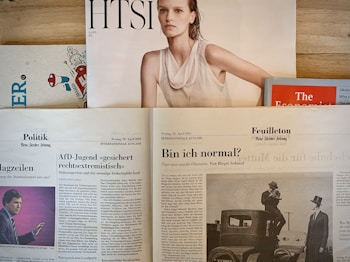 Several newspapers and magazines are arranged on a wooden surface. One magazine features a portrait of a woman with wet hair in a white top. Below are pages from a German newspaper with articles and images, including one of a man in a suit. The Economist magazine is partially visible on the right.