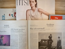 Several newspapers and magazines are arranged on a wooden surface. One magazine features a portrait of a woman with wet hair in a white top. Below are pages from a German newspaper with articles and images, including one of a man in a suit. The Economist magazine is partially visible on the right.
