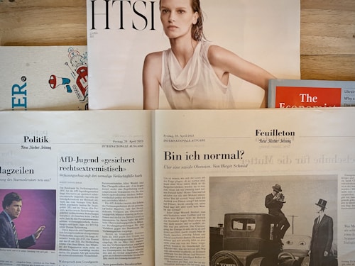 Several newspapers and magazines are arranged on a wooden surface. One magazine features a portrait of a woman with wet hair in a white top. Below are pages from a German newspaper with articles and images, including one of a man in a suit. The Economist magazine is partially visible on the right.