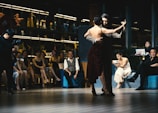 A couple is performing a tango dance in front of a seated audience. The dancers are elegantly dressed, with the woman wearing a dark, sparkling dress. The audience appears engaged and entertained, with some people capturing moments with their phones. The room is dimly lit, creating an intimate atmosphere, and decorated with various figurines and potted plants.