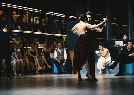 A couple is performing a tango dance in front of a seated audience. The dancers are elegantly dressed, with the woman wearing a dark, sparkling dress. The audience appears engaged and entertained, with some people capturing moments with their phones. The room is dimly lit, creating an intimate atmosphere, and decorated with various figurines and potted plants.