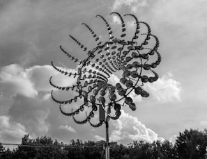 A large, intricate metal sculpture with a spiral design stands against a backdrop of a cloudy sky. The sculpture consists of numerous reflective discs that create a mesmerizing pattern, reminiscent of a wind sculpture or kinetic art. Below the sculpture, there is a line of trees partially visible.