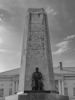 A tall stone monument with intricate relief carvings depicting historical scenes. At the base, there is a seated statue of a person, hands resting on their lap. The architecture behind the monument includes classical columns and large windows. The sky above is partly cloudy, adding texture to the backdrop.