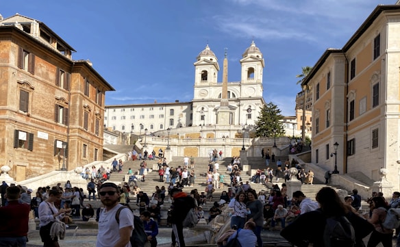 A bustling tourist area with a large staircase leading up to a historic church featuring twin towers. The space is surrounded by old buildings with distinct architecture and filled with people interacting, taking photos, and relaxing under a clear blue sky.