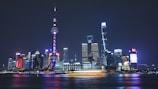 Bright, illuminated skyscrapers create a stunning skyline at night, with reflections on the water in the foreground. The iconic Oriental Pearl Tower stands prominently among other modern buildings, glowing with vibrant colors.