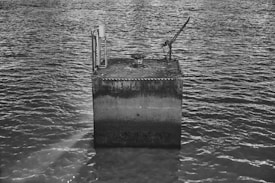 A concrete structure protruding from the water, equipped with a metallic platform and a mechanical apparatus. The water surface is slightly rippled, creating a serene texture across the frame. The image is in monochrome, enhancing the texture and details of the scene.