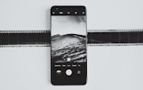 A smartphone displays a black and white photograph of a landscape with clouds and hills. The phone is placed on top of a film strip, creating a contrast between modern and traditional photography methods.