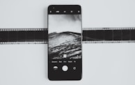 A smartphone displays a black and white photograph of a landscape with clouds and hills. The phone is placed on top of a film strip, creating a contrast between modern and traditional photography methods.