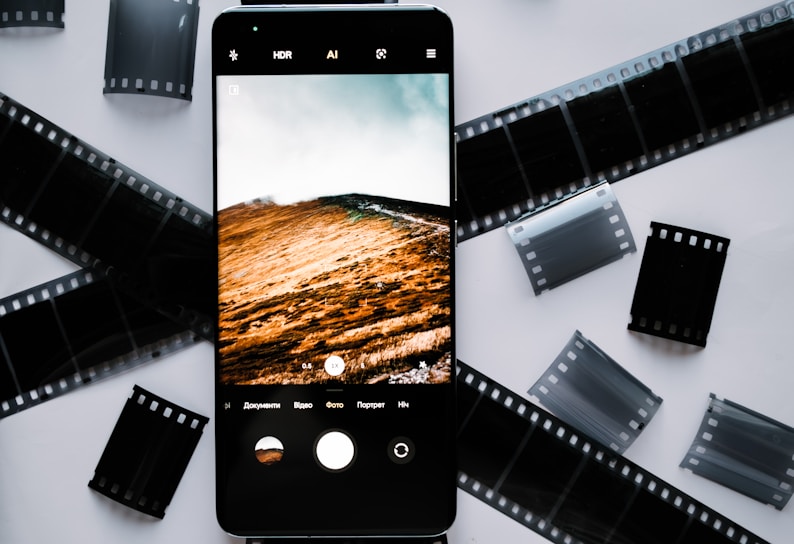 A smartphone lies on a white surface surrounded by several black and transparent film strips. The phone's screen displays a vivid landscape of hills and a cloudy sky, captured through its camera app.
