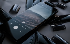 A smartphone displaying a photo or camera app is laid on a surface surrounded by several strips of camera film. The phone screen shows a landscape image, while the surrounding film strips are reminiscent of traditional photography.