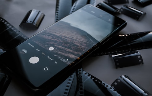 A smartphone displaying a photo or camera app is laid on a surface surrounded by several strips of camera film. The phone screen shows a landscape image, while the surrounding film strips are reminiscent of traditional photography.
