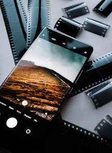 A smartphone displaying a photo of a landscape with rolling hills and a cloudy sky is placed on top of several film negatives scattered on a table. The screen of the smartphone shows camera interface icons, indicating it is in photography mode.