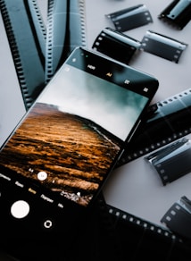 A smartphone displaying a photo of a landscape with rolling hills and a cloudy sky is placed on top of several film negatives scattered on a table. The screen of the smartphone shows camera interface icons, indicating it is in photography mode.