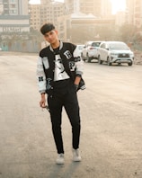 A young person is standing in a casual pose on an urban street. They are wearing a black and white varsity jacket, a T-shirt, black pants, and white sneakers. The individual is holding a pair of sunglasses. In the background, there are several parked cars and tall buildings, with the sun casting a warm glow.