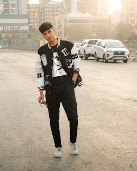 A young person is standing in a casual pose on an urban street. They are wearing a black and white varsity jacket, a T-shirt, black pants, and white sneakers. The individual is holding a pair of sunglasses. In the background, there are several parked cars and tall buildings, with the sun casting a warm glow.