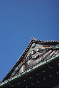 A traditional architectural roof detail with ornate woodwork and intricate designs against a clear blue sky. The roof features decorative ridge caps and elaborate carvings that suggest cultural or historical significance.