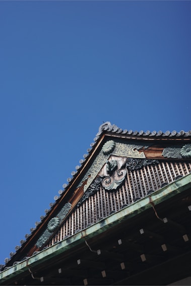 A traditional architectural roof detail with ornate woodwork and intricate designs against a clear blue sky. The roof features decorative ridge caps and elaborate carvings that suggest cultural or historical significance.