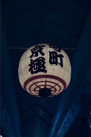 A large traditional Japanese lantern with kanji characters hangs in an indoor setting with an arching ceiling. The lantern is illuminated, casting a warm glow, while the background remains dimly lit.