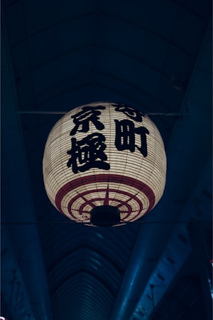 A large traditional Japanese lantern with kanji characters hangs in an indoor setting with an arching ceiling. The lantern is illuminated, casting a warm glow, while the background remains dimly lit.