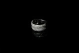 A silver ring encrusted with small sparkling stones is set against a black background, creating a striking contrast. The reflective surface beneath the ring enhances its shine and adds depth to the image.