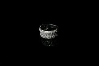 A silver ring encrusted with small sparkling stones is set against a black background, creating a striking contrast. The reflective surface beneath the ring enhances its shine and adds depth to the image.