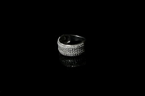 A silver ring encrusted with small sparkling stones is set against a black background, creating a striking contrast. The reflective surface beneath the ring enhances its shine and adds depth to the image.