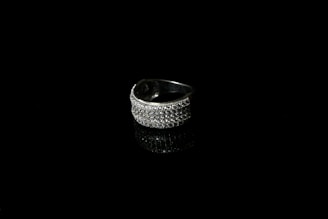 A silver ring encrusted with small sparkling stones is set against a black background, creating a striking contrast. The reflective surface beneath the ring enhances its shine and adds depth to the image.