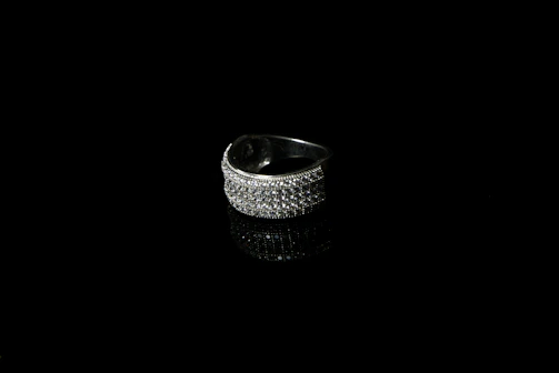 A silver ring encrusted with small sparkling stones is set against a black background, creating a striking contrast. The reflective surface beneath the ring enhances its shine and adds depth to the image.