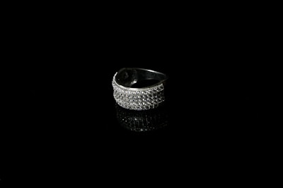 A silver ring encrusted with small sparkling stones is set against a black background, creating a striking contrast. The reflective surface beneath the ring enhances its shine and adds depth to the image.