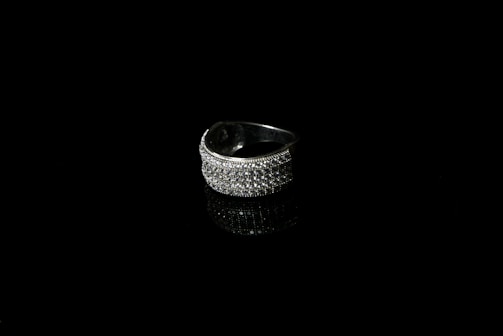 A silver ring encrusted with small sparkling stones is set against a black background, creating a striking contrast. The reflective surface beneath the ring enhances its shine and adds depth to the image.
