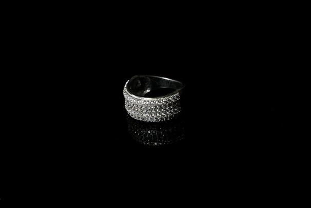 A silver ring encrusted with small sparkling stones is set against a black background, creating a striking contrast. The reflective surface beneath the ring enhances its shine and adds depth to the image.
