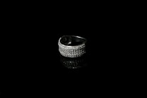 A silver ring encrusted with small sparkling stones is set against a black background, creating a striking contrast. The reflective surface beneath the ring enhances its shine and adds depth to the image.