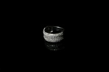 A silver ring encrusted with small sparkling stones is set against a black background, creating a striking contrast. The reflective surface beneath the ring enhances its shine and adds depth to the image.